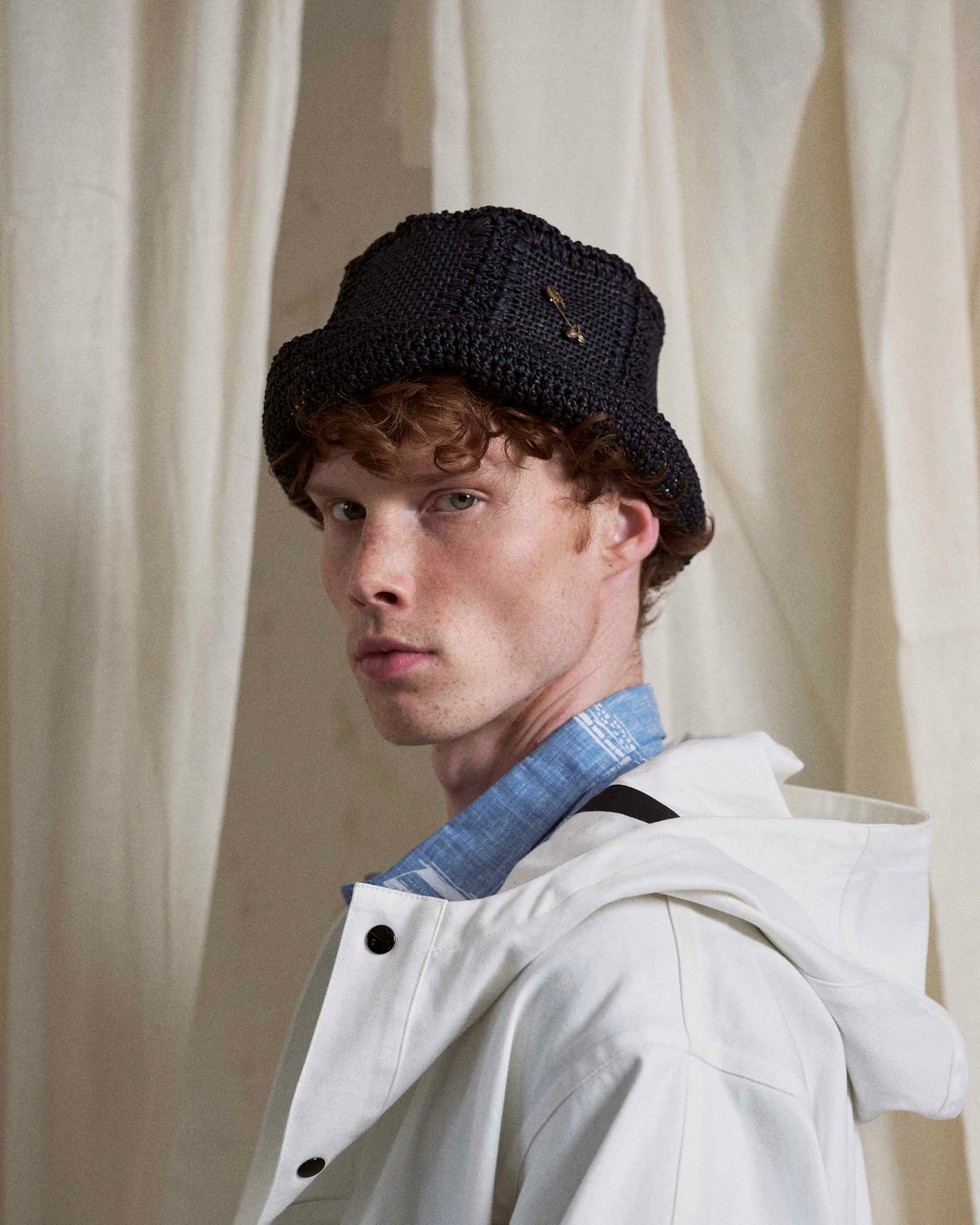 Man wearing a handmade black woven hat and white coat against a neutral fabric background