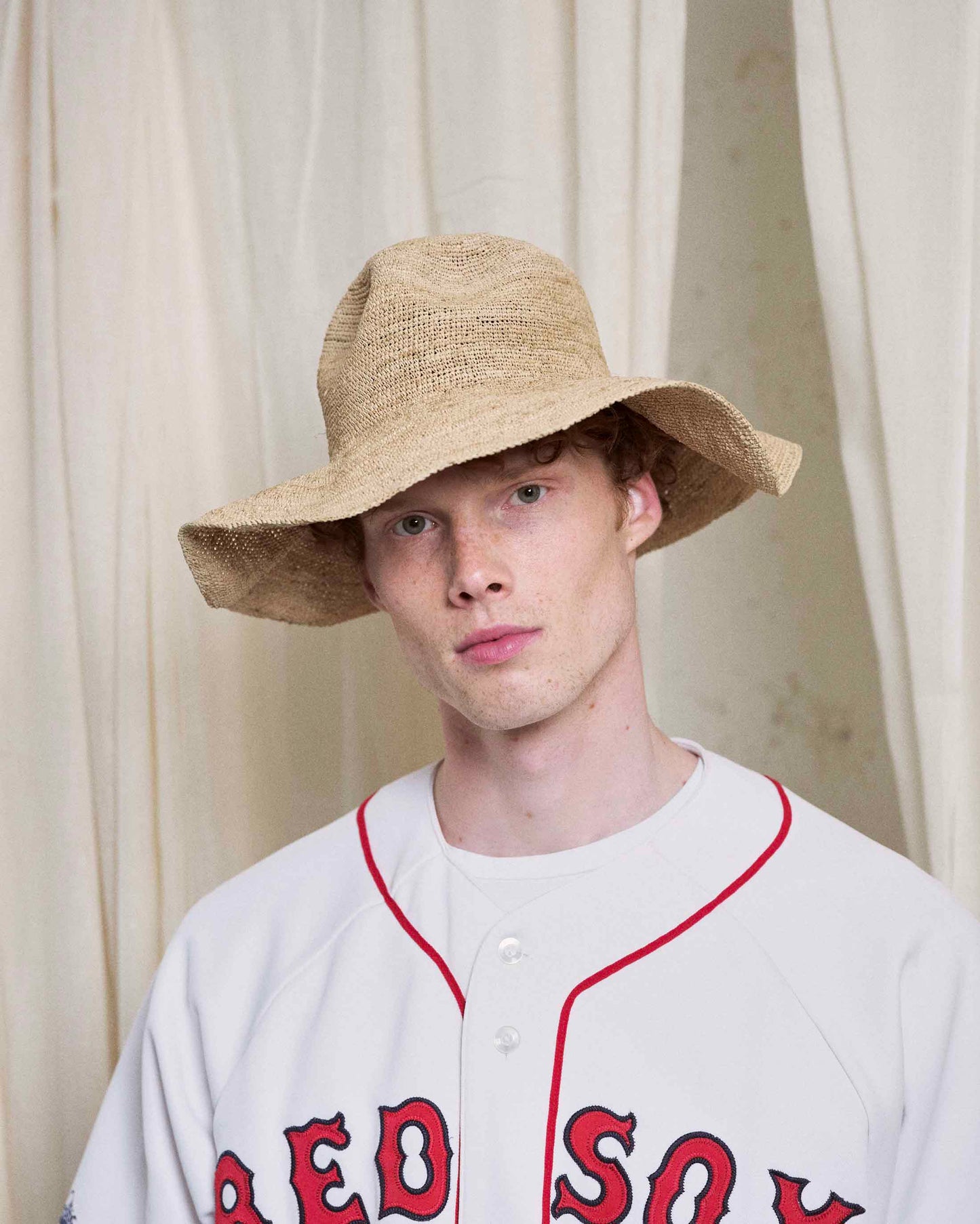 Man in a wide-brimmed straw hat and white baseball jersey, standing by cream curtains.