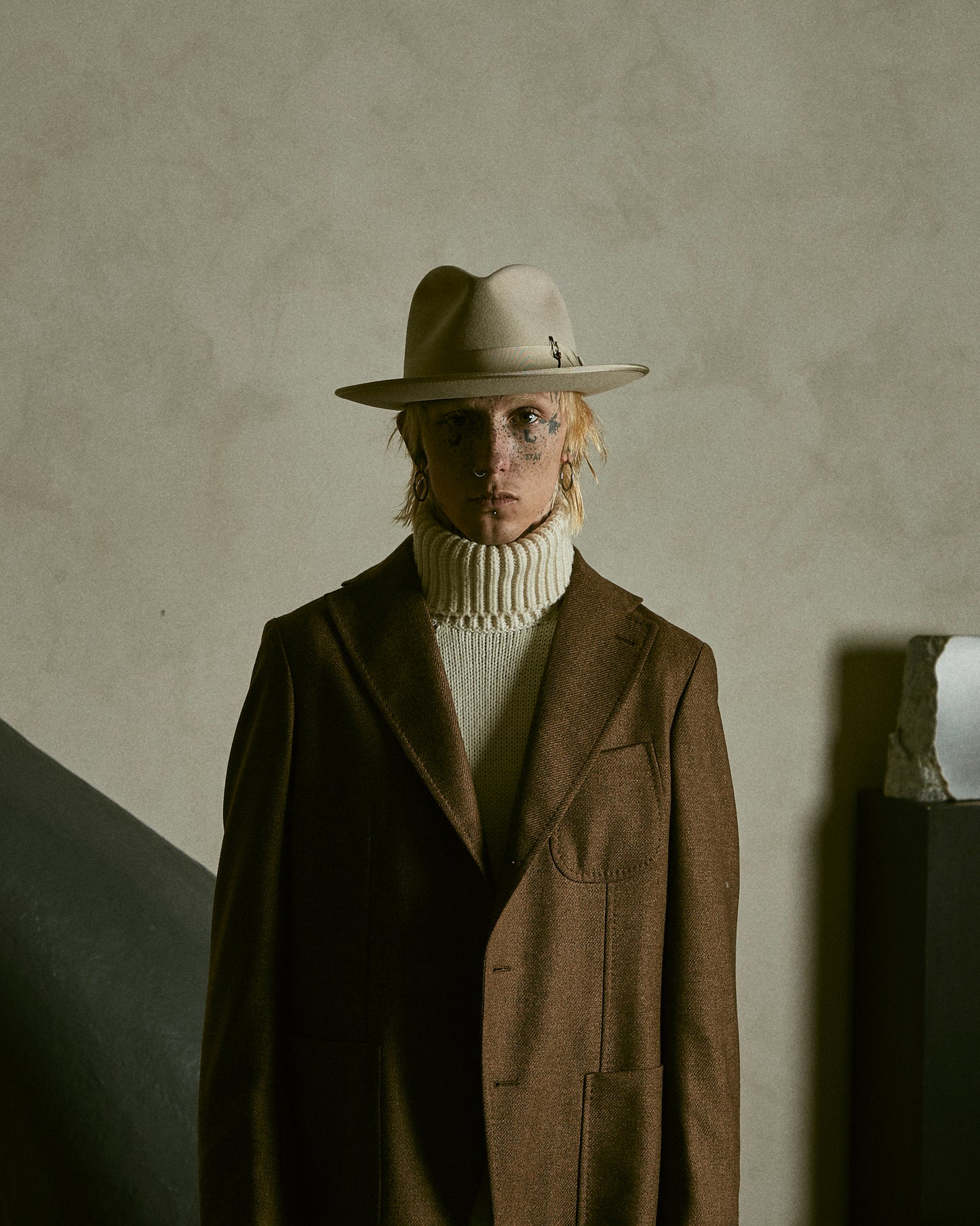Person wearing a cream felt hat, white turtleneck sweater, and brown blazer in a studio setting.