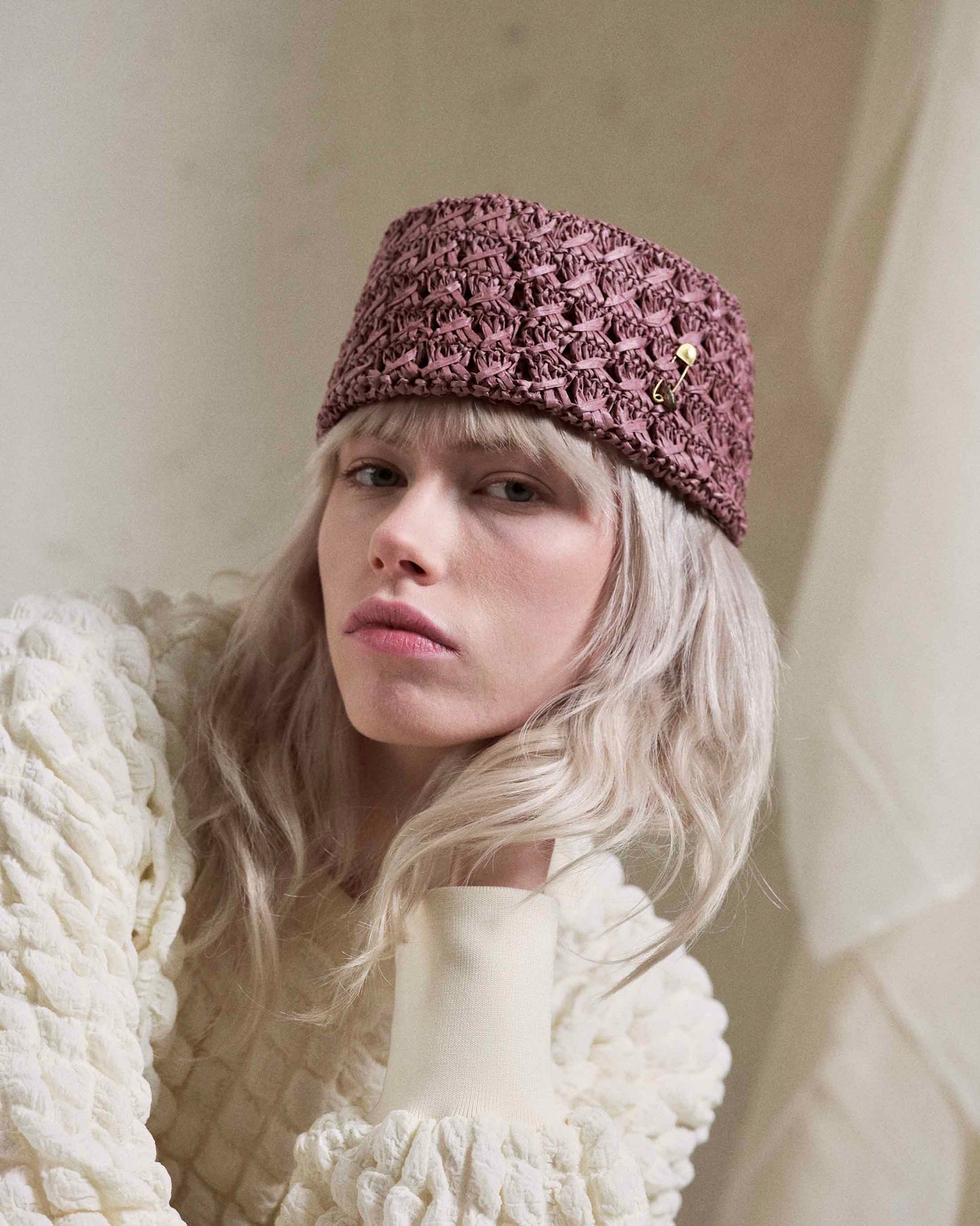 Woman wearing a handmade pink woven hat with gold pin, textured cream sweater, neutral background.