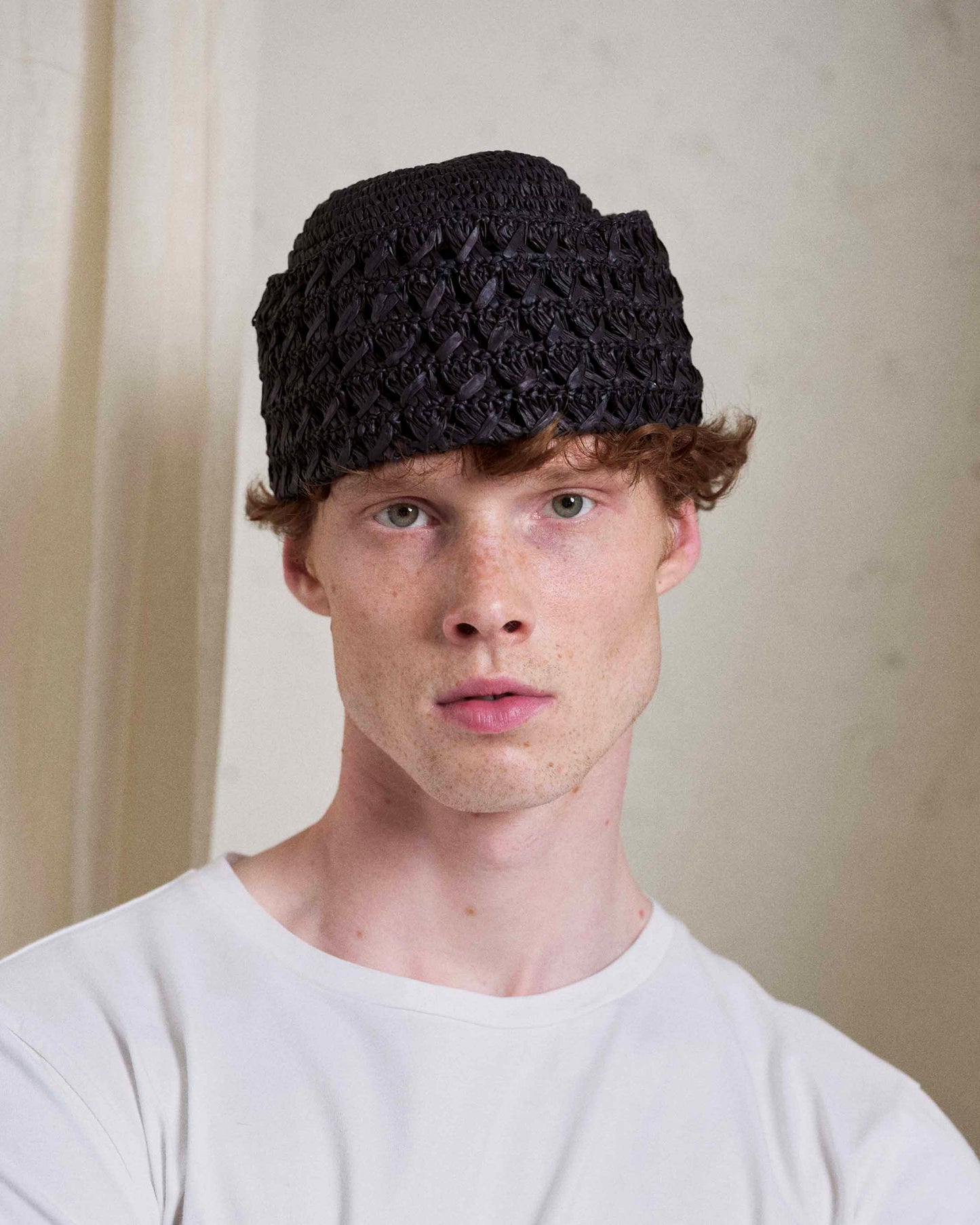Man wearing black woven Italian hat, casual white shirt, minimalist neutral background