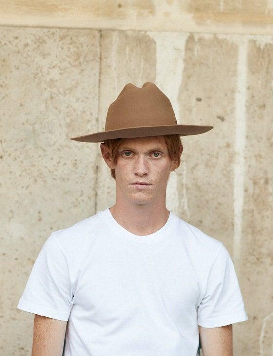 Man wearing a wide-brimmed brown felt hat and white t-shirt against a stone wall background