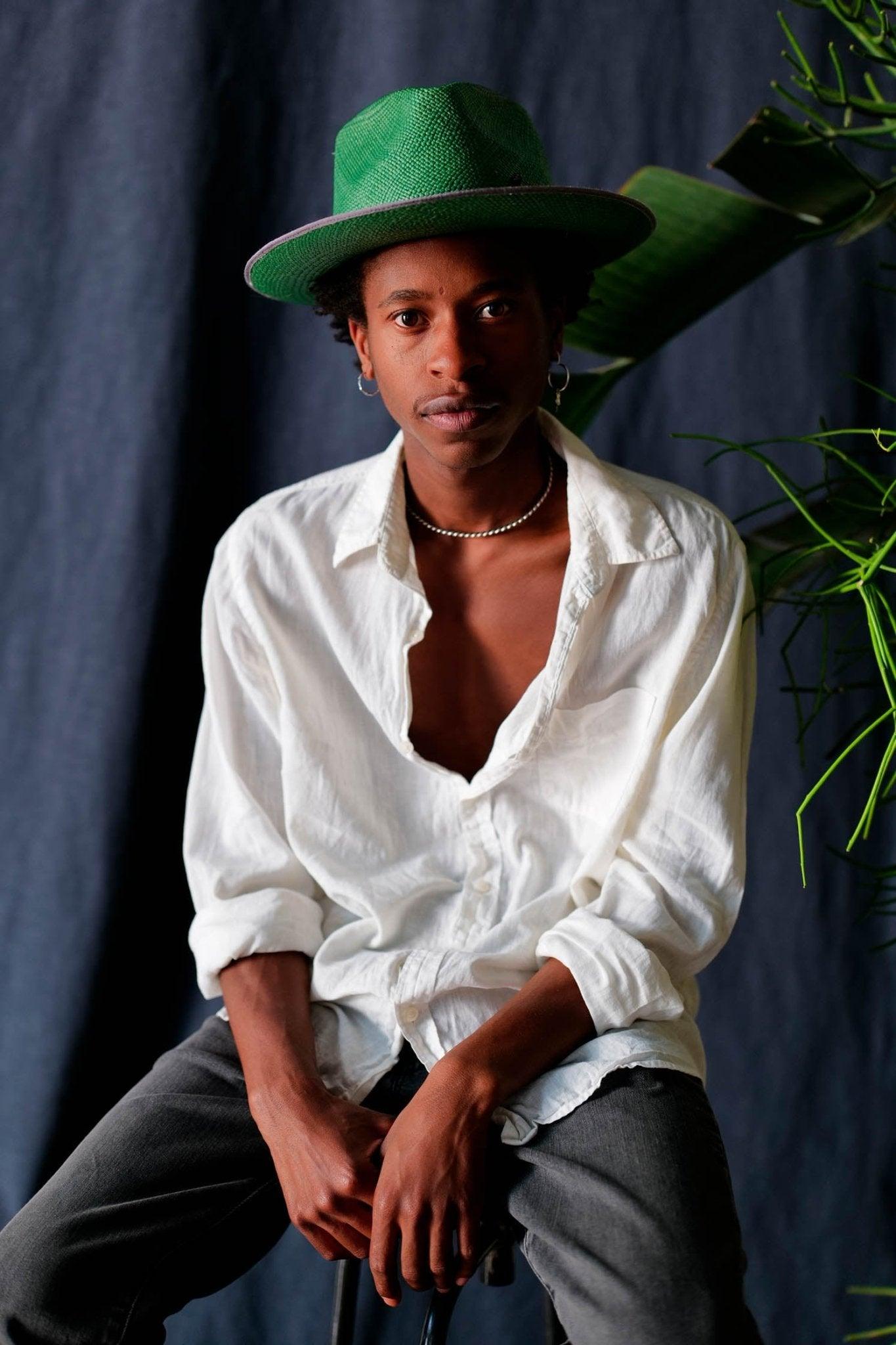 Man wearing a green Panama hat, white shirt, and dark pants against a dark background with plants.