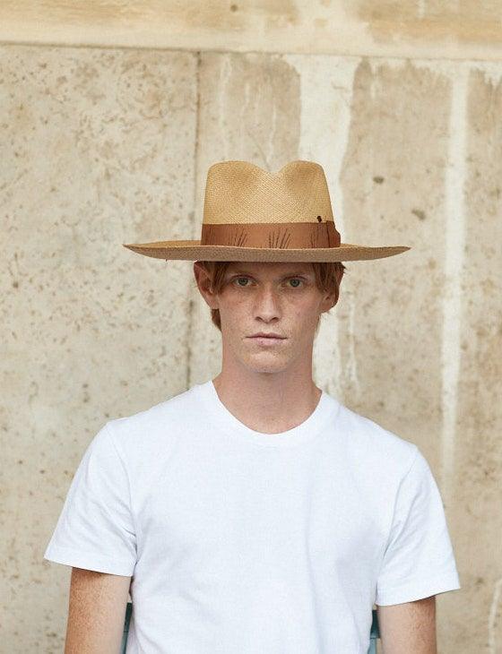 Man wearing a wide-brim khaki straw hat with brown band, white shirt, stone wall background