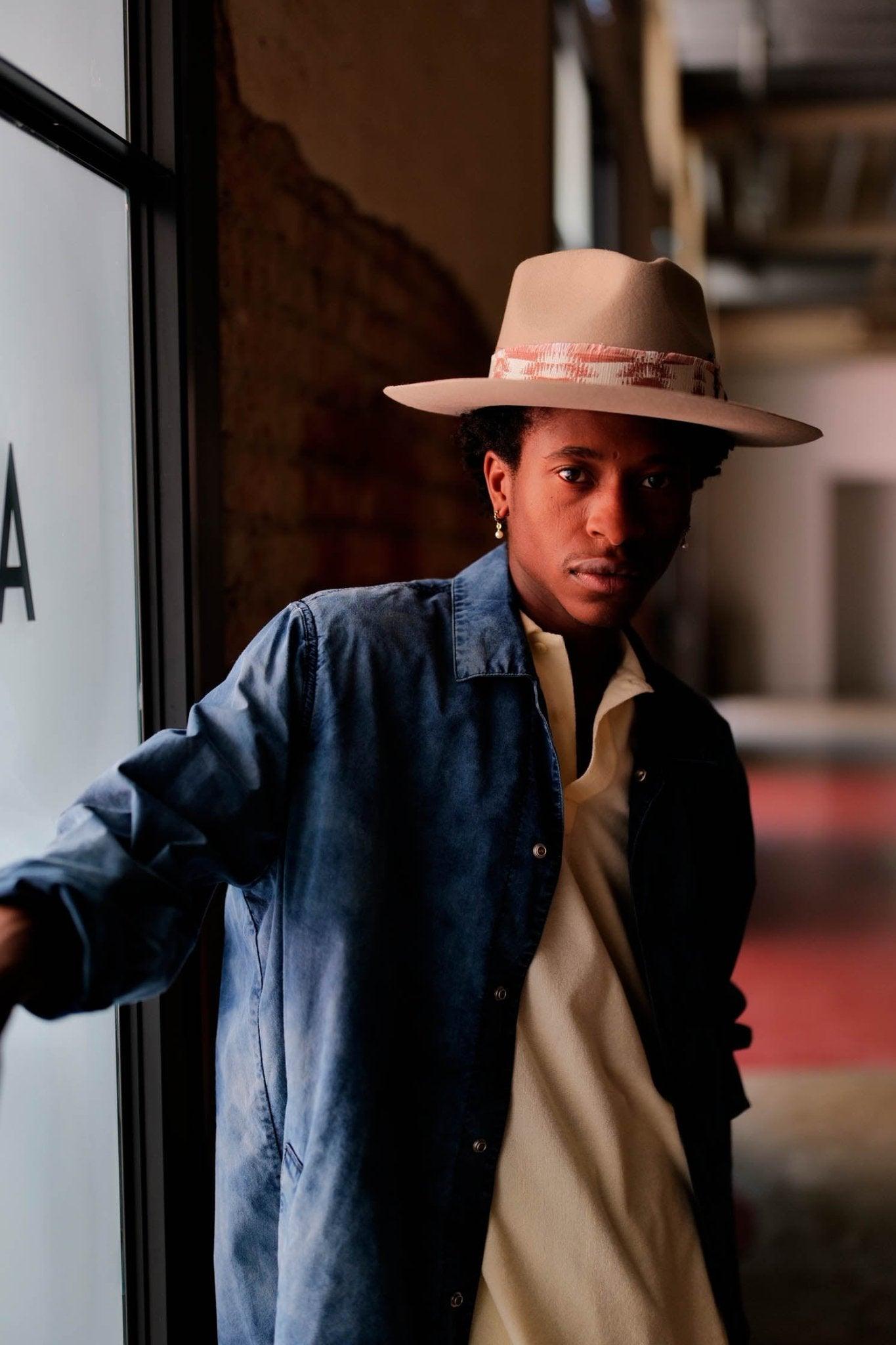 Man wearing a light tan felt hat with patterned band, blue jacket, and cream shirt indoors