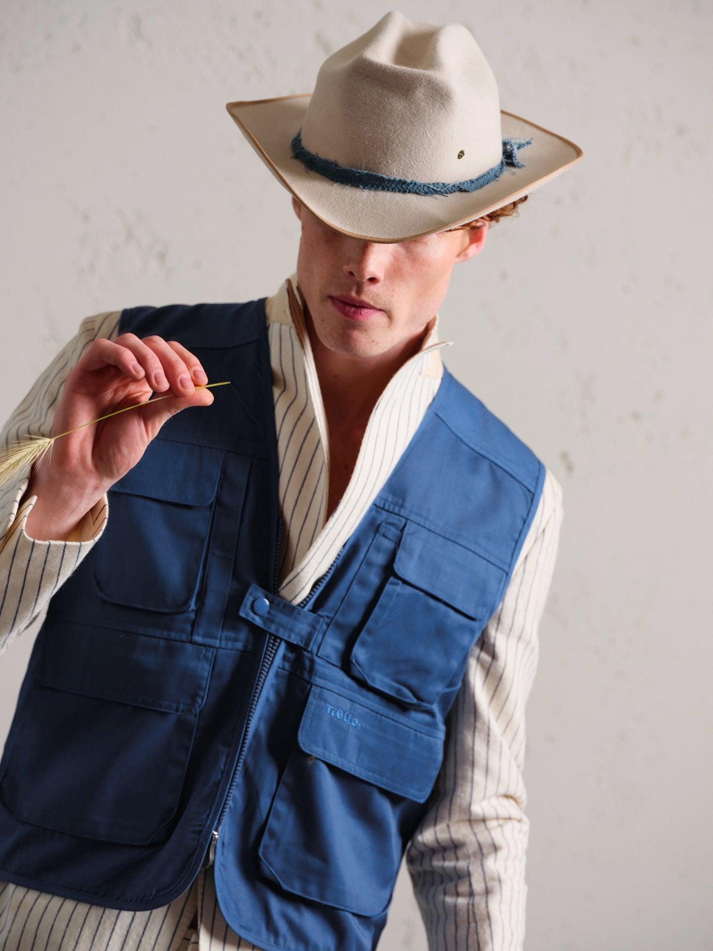Man wearing a cream felt hat with blue trim and a blue utility vest over a striped shirt