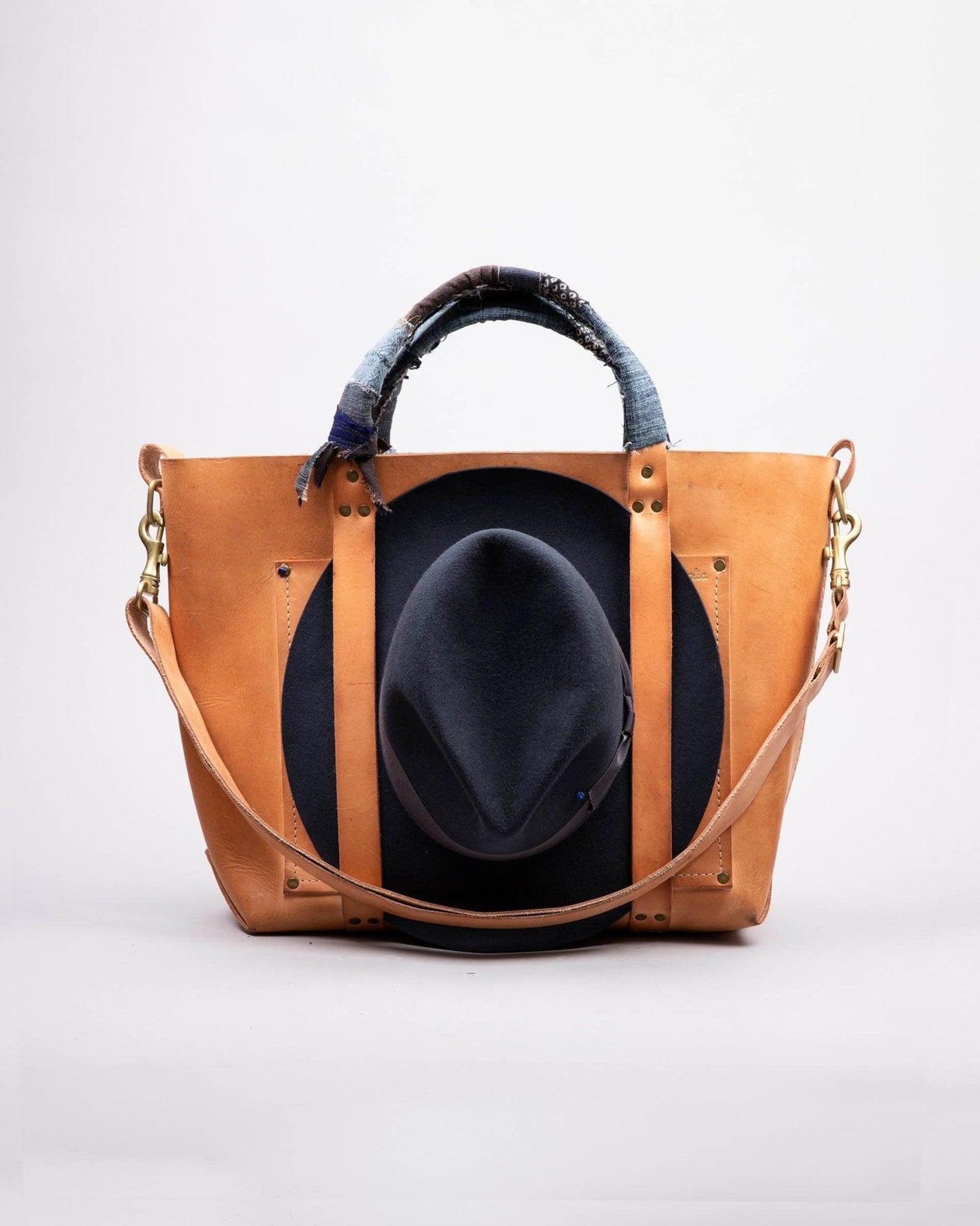 Tan leather tote bag with denim-wrapped handles and a dark felt fedora hat inside